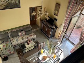 A living room with a sofa, a glass coffee table, and a dining table with chairs. There is a television on a cabinet, a chandelier, and a window with curtains.