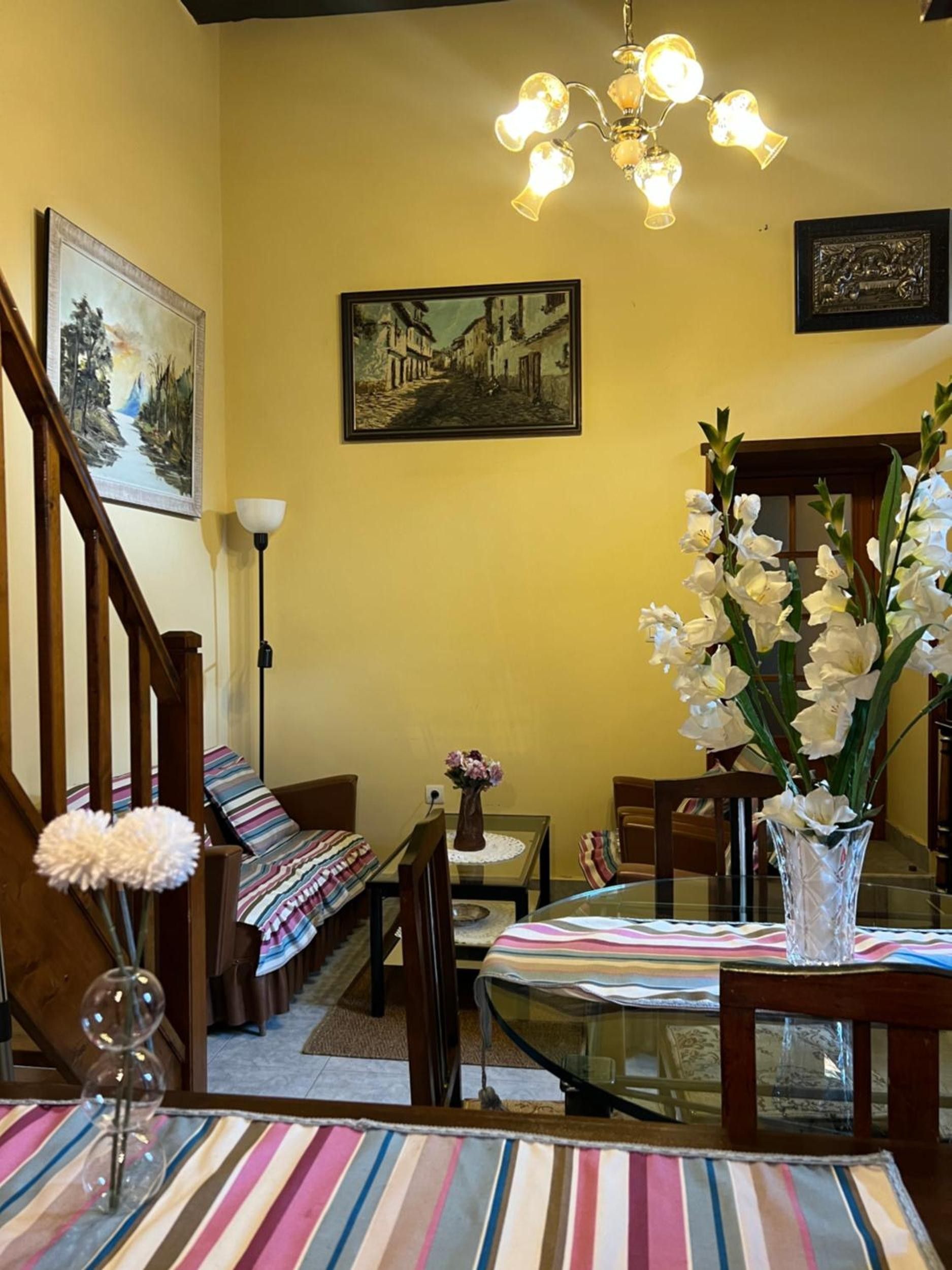 Un coin repas avec une table en verre, une nappe à rayures, des chaises, un vase de fleurs, un tableau et un lustre.