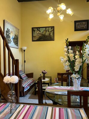 A dining area with a glass table, striped tablecloth, chairs, a vase with flowers, a painting, and a chandelier.