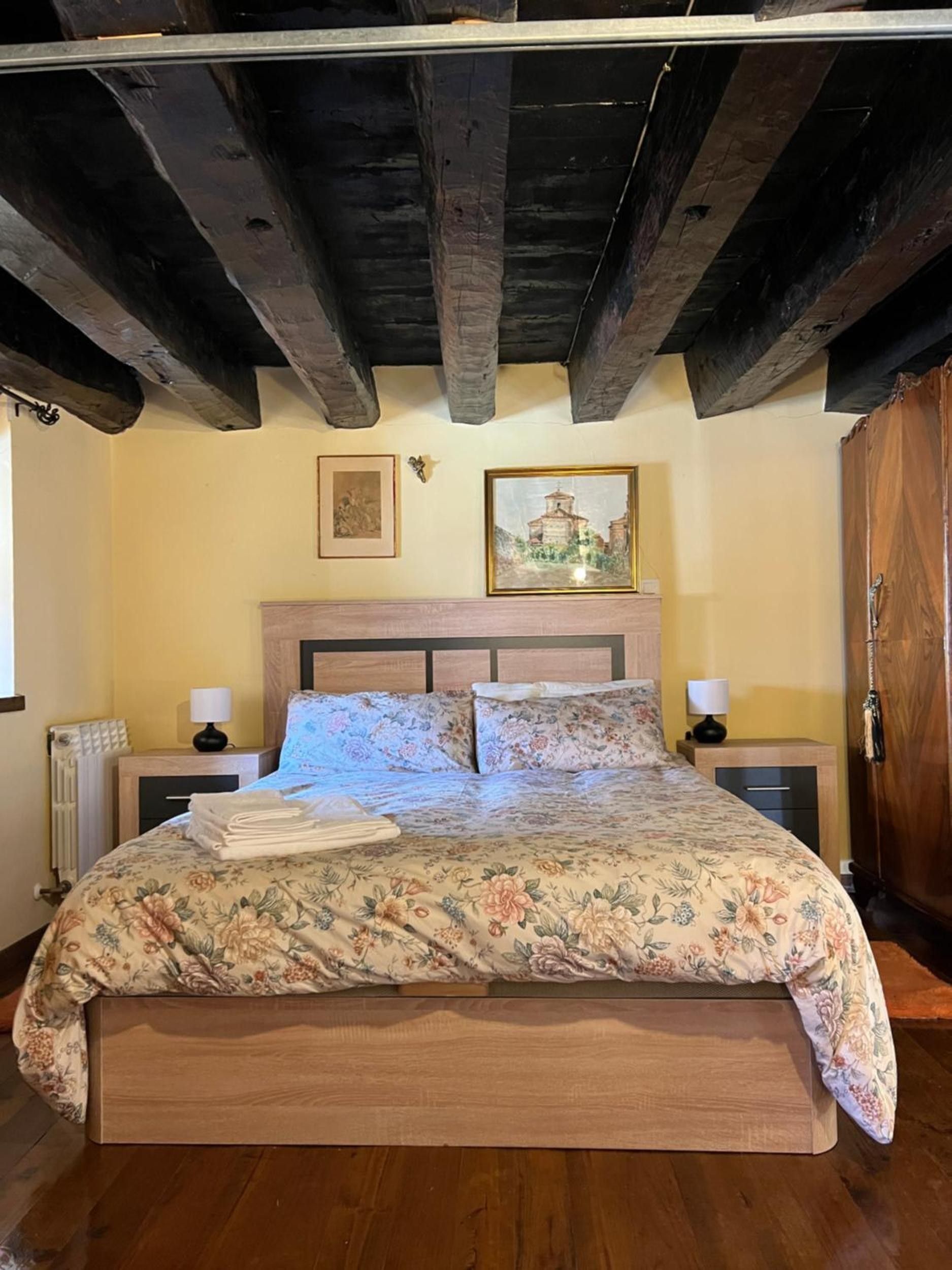 A bedroom with a wooden headboard, a floral bedspread, two bedside tables with lamps, and a wooden wardrobe.