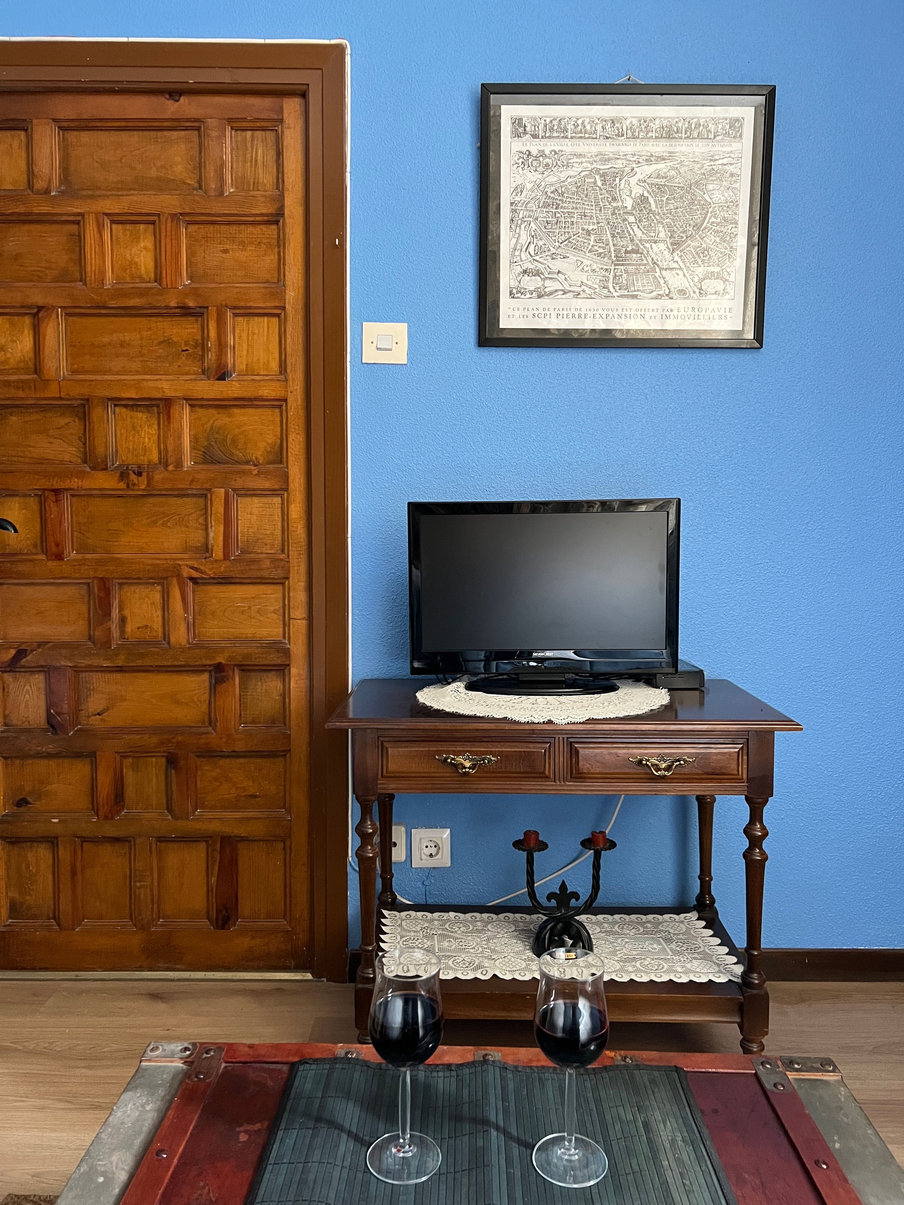 Ein Zimmer mit einer Holztür, einer gerahmten Karte an der Wand, einem Fernseher auf einem Holztisch und zwei Weingläsern auf einer Matte.