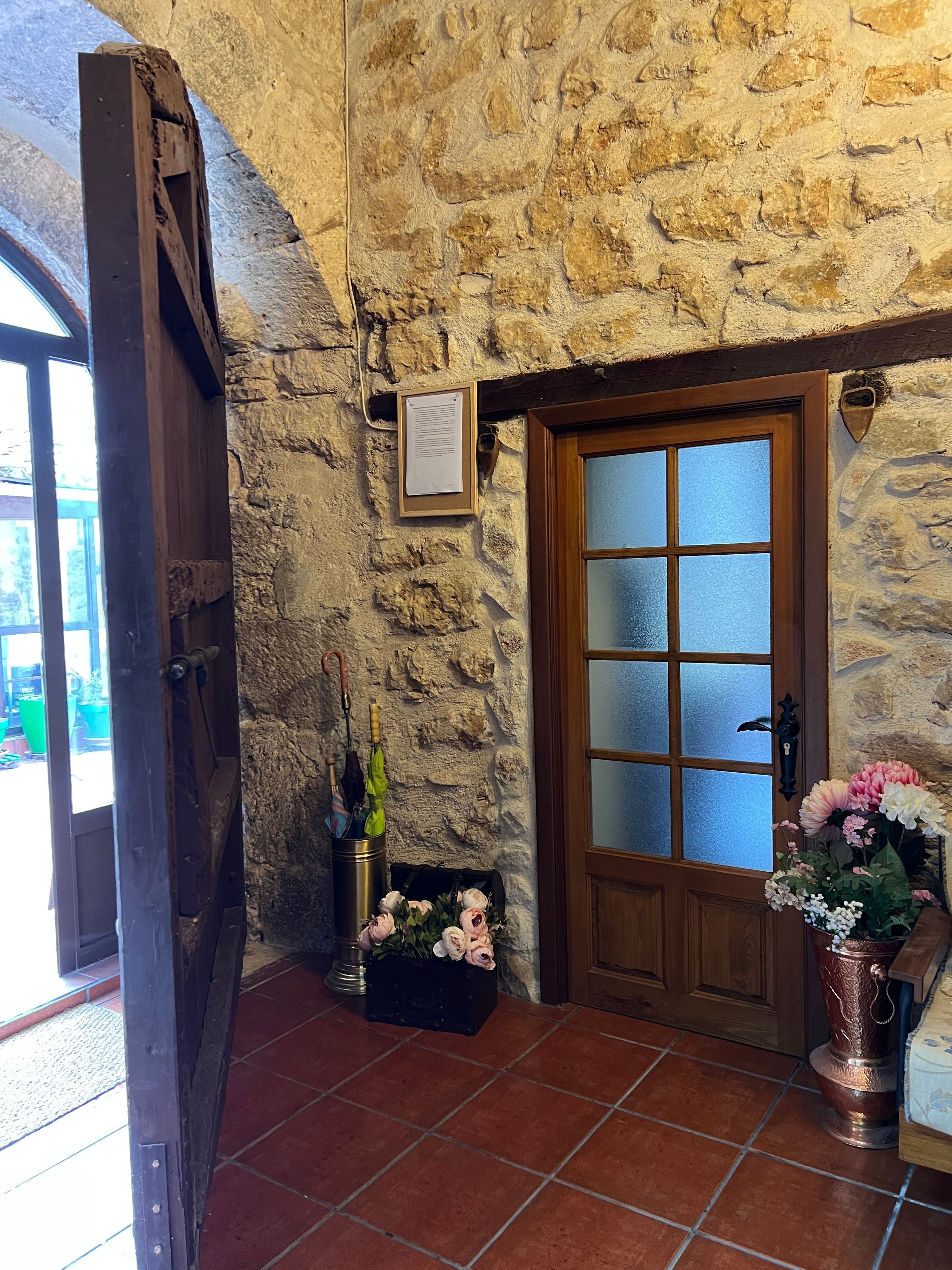 Un mur de pierre, une porte en bois avec des panneaux de verre dépoli, un seau en métal rempli de fleurs et un autre seau en métal contenant des parapluies.