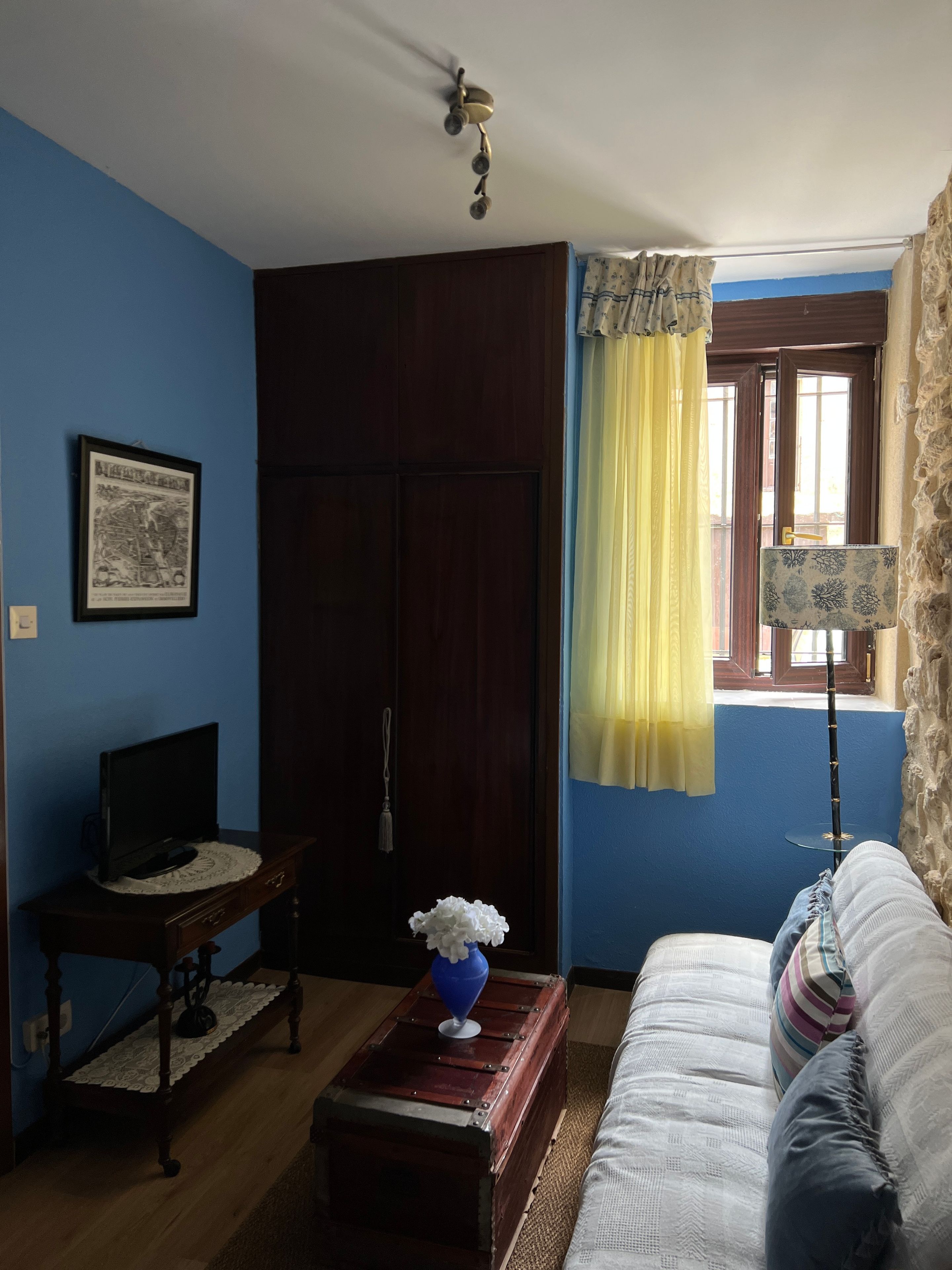 Une pièce avec une armoire en bois foncé, un téléviseur posé sur une petite table, un canapé avec des coussins, une fenêtre avec des rideaux et une lampe.