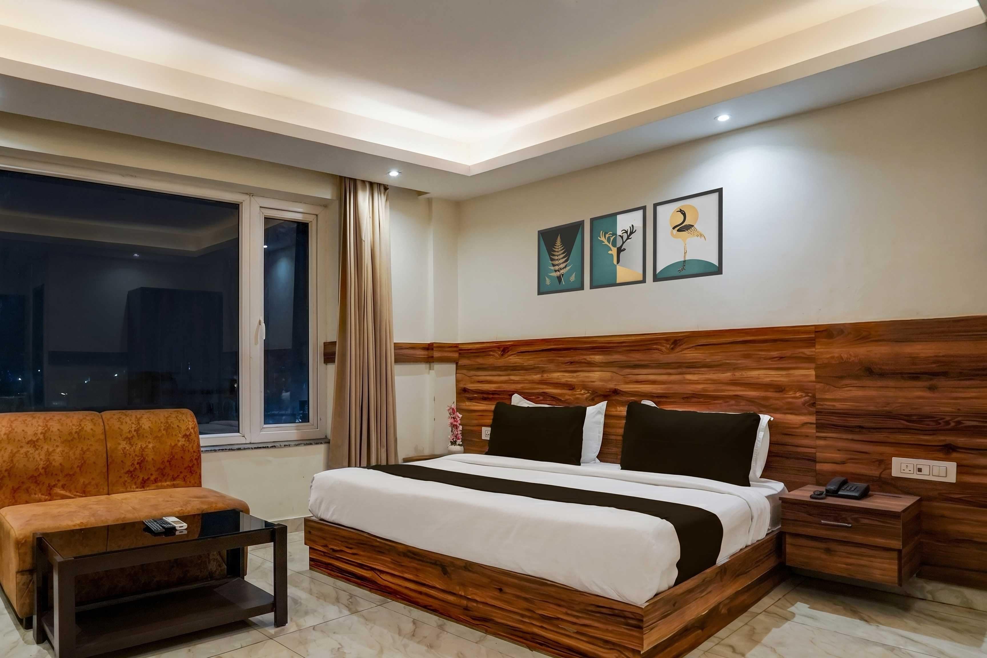 A hotel room with a bed, a nightstand, a small table, a sofa, a window with curtains, and wall art.