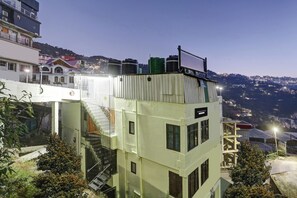 Un edificio de varios pisos con una escalera, una azotea con tanques de agua y una vista de un pueblo montañoso al fondo.