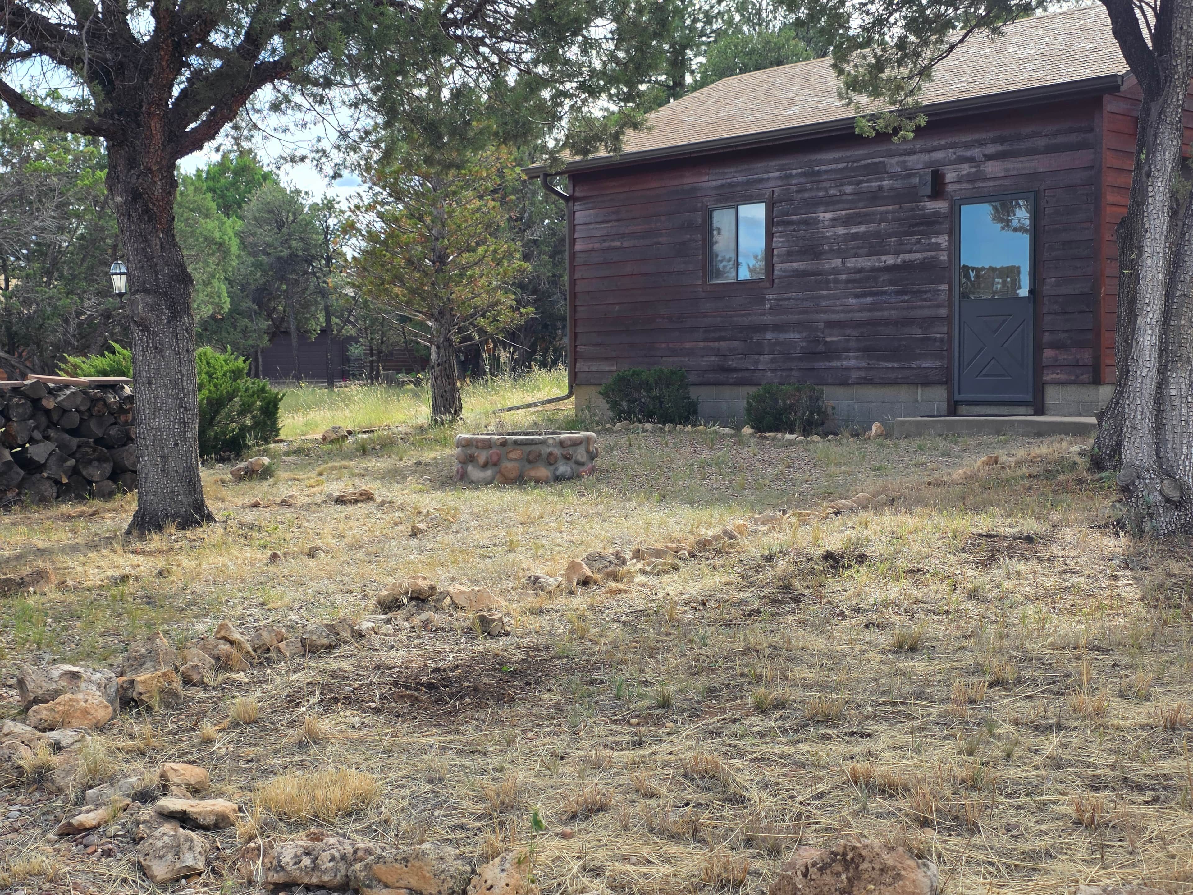 A rustic wooden cabin with a stone fire pit, surrounded by dry grass and trees.