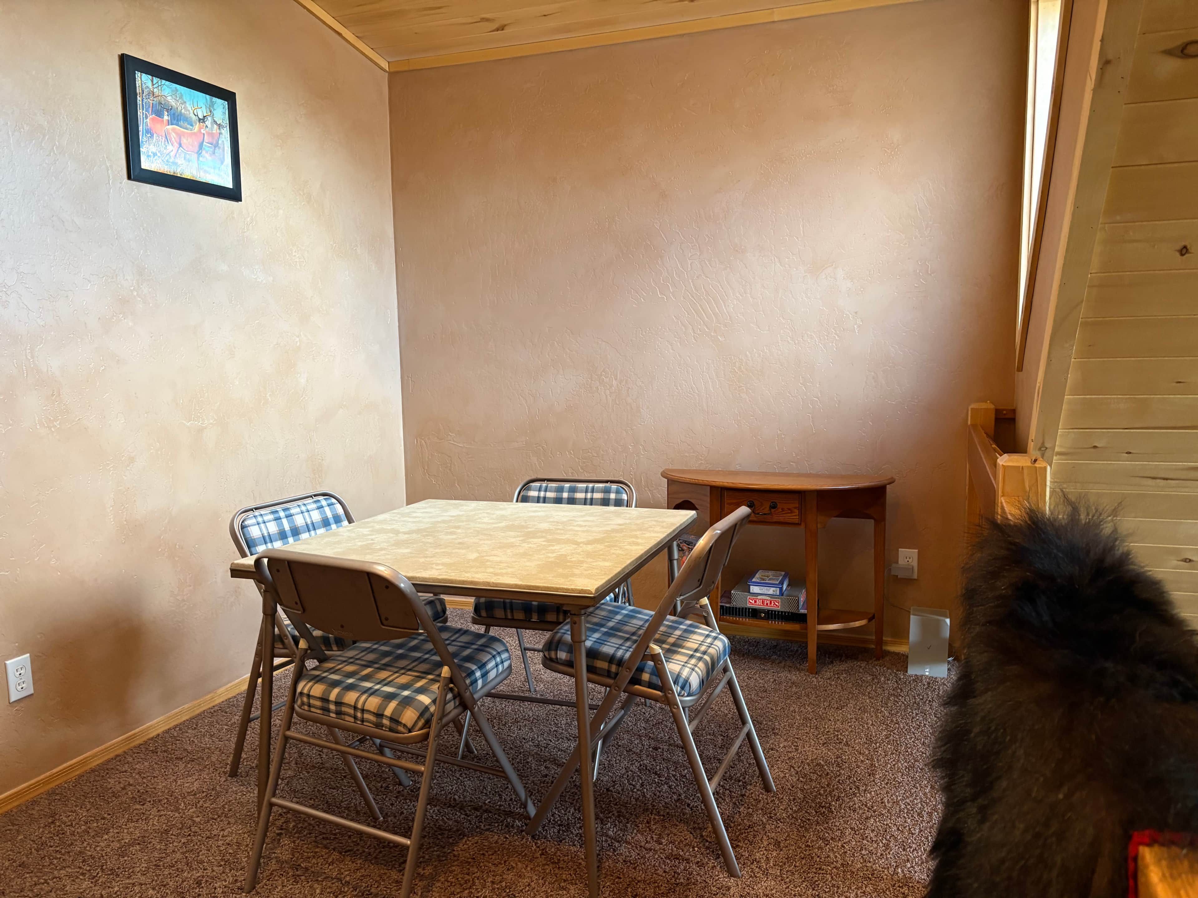 A room with a wooden table, four chairs with plaid cushions, a small wooden side table, and a framed picture on the wall.