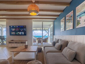 A living room with a TV, a wooden table, a sofa, and a view of the ocean.