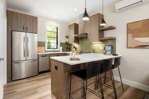 Eine moderne Küche mit Edelstahlkühlschrank, Holzschränken, einer zentralen Kochinsel mit Barhocker und Pendelleuchten.
