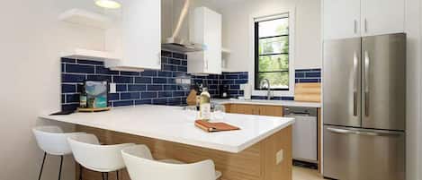 Una cocina moderna con una isla central, taburetes de bar, electrodomésticos de acero inoxidable y un ventanal que proporciona luz natural.