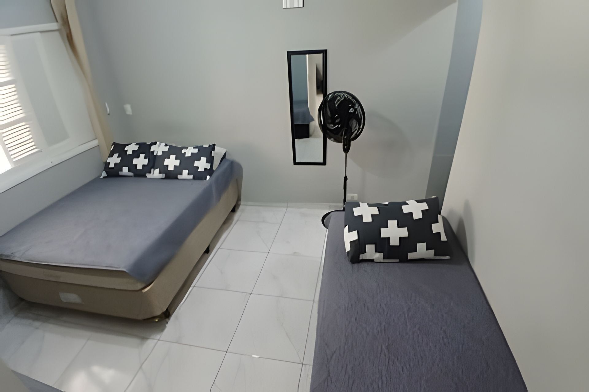 Um quarto pequeno e simples com duas camas, um ventilador e um espelho na parede.