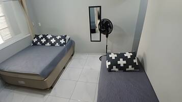 Um quarto pequeno e simples com duas camas, um ventilador e um espelho na parede.