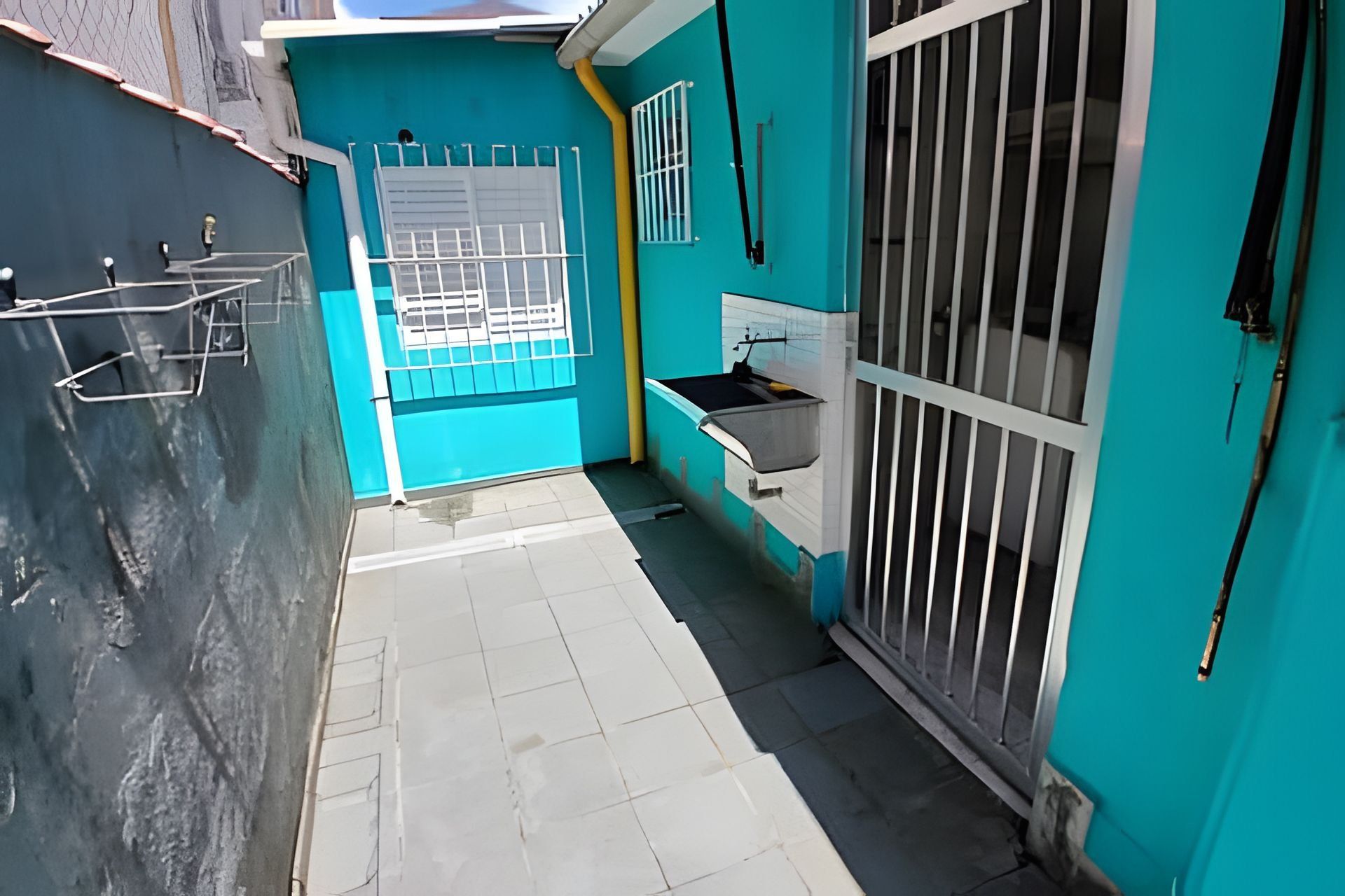 Un espacio exterior pequeño y cercado, con suelo de baldosas, una puerta metálica, un lavamanos y una ventana con barrotes.