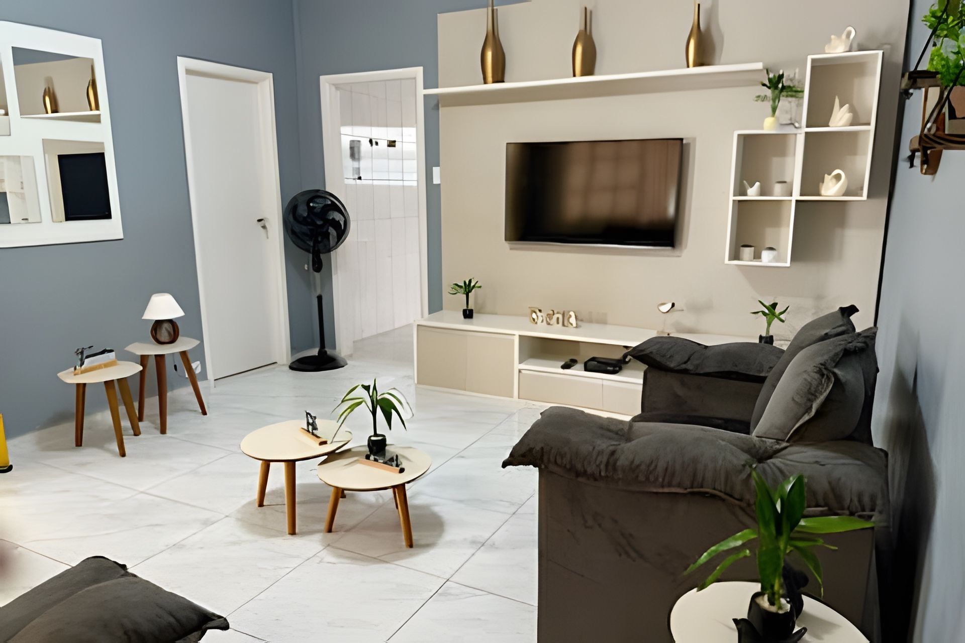 Ein modernes Wohnzimmer mit einem grauen Sofa, einem an der Wand montierten Fernseher und einer weißen Unterhaltungsecke mit Regalen. Zwei kleine runde Tische mit Lampen und ein Standventilator sind ebenfalls vorhanden.