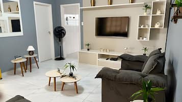 Ein modernes Wohnzimmer mit einem grauen Sofa, einem an der Wand montierten Fernseher und einer weißen Unterhaltungsecke mit Regalen. Zwei kleine runde Tische mit Lampen und ein Standventilator sind ebenfalls vorhanden.