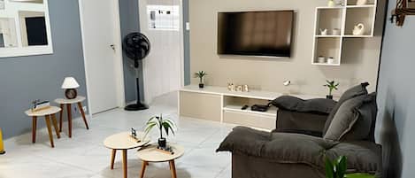 Una sala de estar moderna con un sofá gris, un televisor montado en la pared y un mueble blanco para entretenimiento con estantes. Hay dos pequeñas mesas redondas con lámparas y un ventilador de pie.