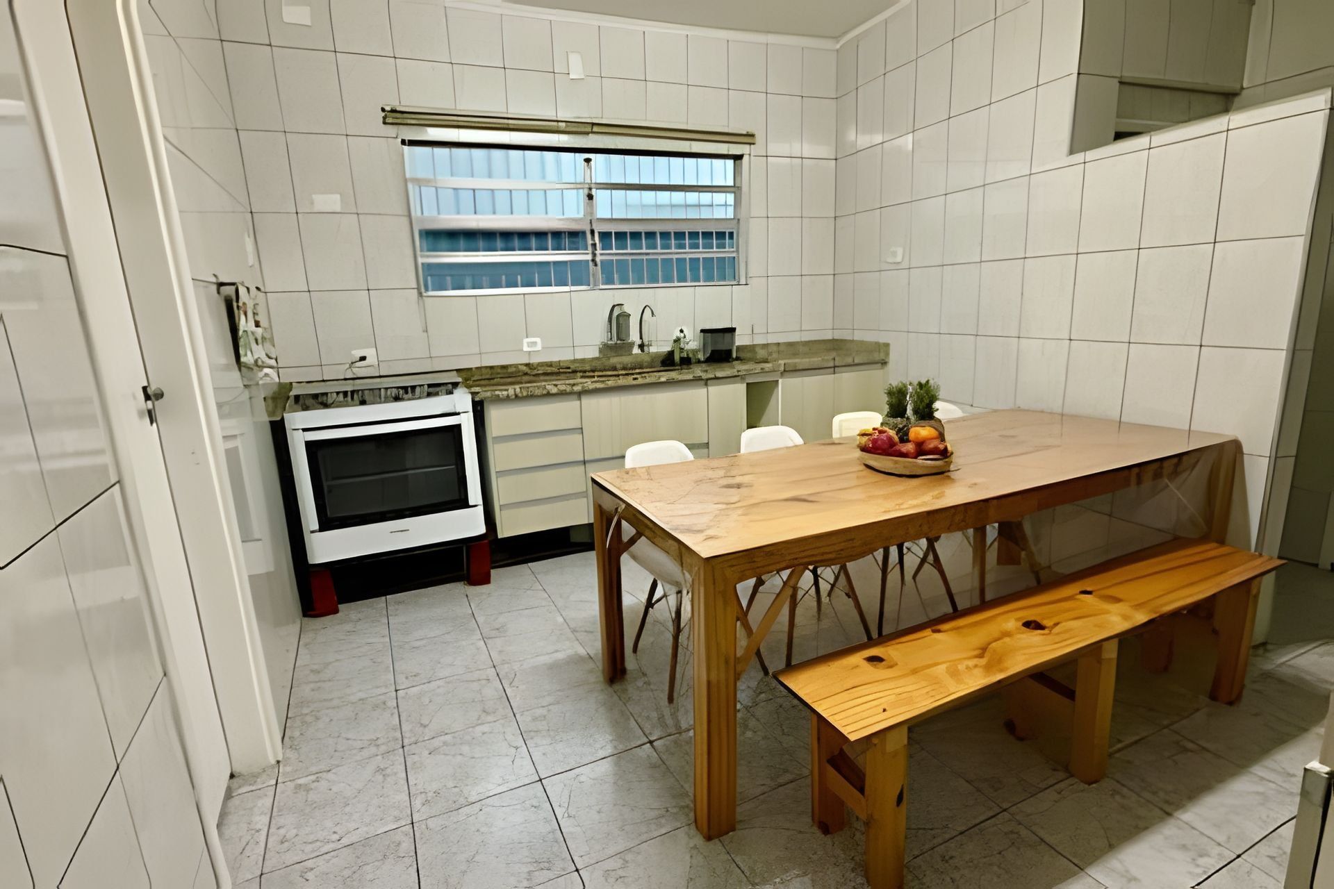 Uma cozinha com azulejos brancos, uma mesa de madeira com um banco e uma fruteira sobre a mesa.