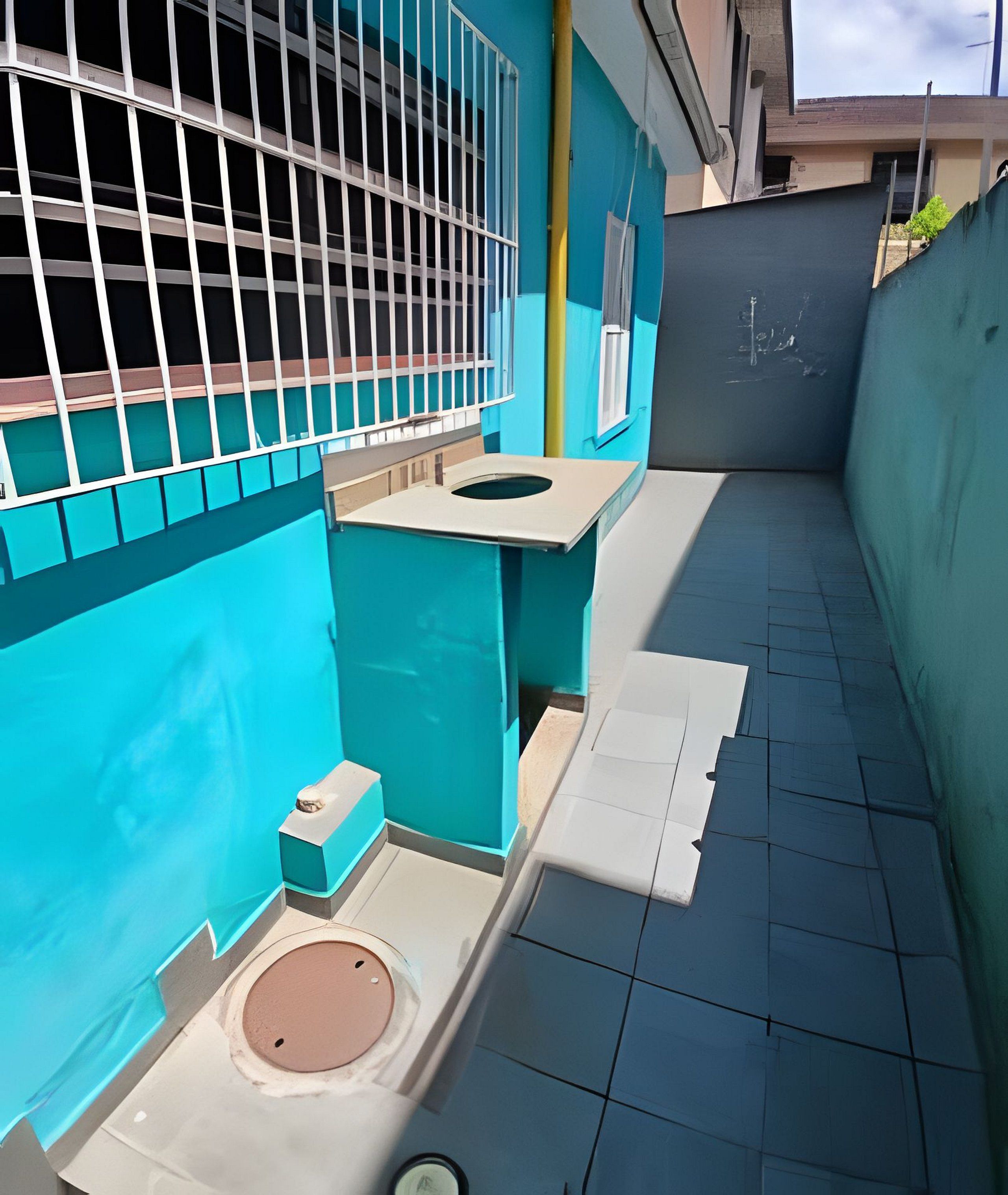 Un estrecho pasillo exterior con una pared azul, una reja metálica y una pequeña mesa blanca con un taburete.