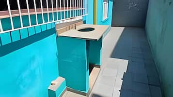 Un estrecho pasillo exterior con una pared azul, una reja metálica y una pequeña mesa blanca con un taburete.
