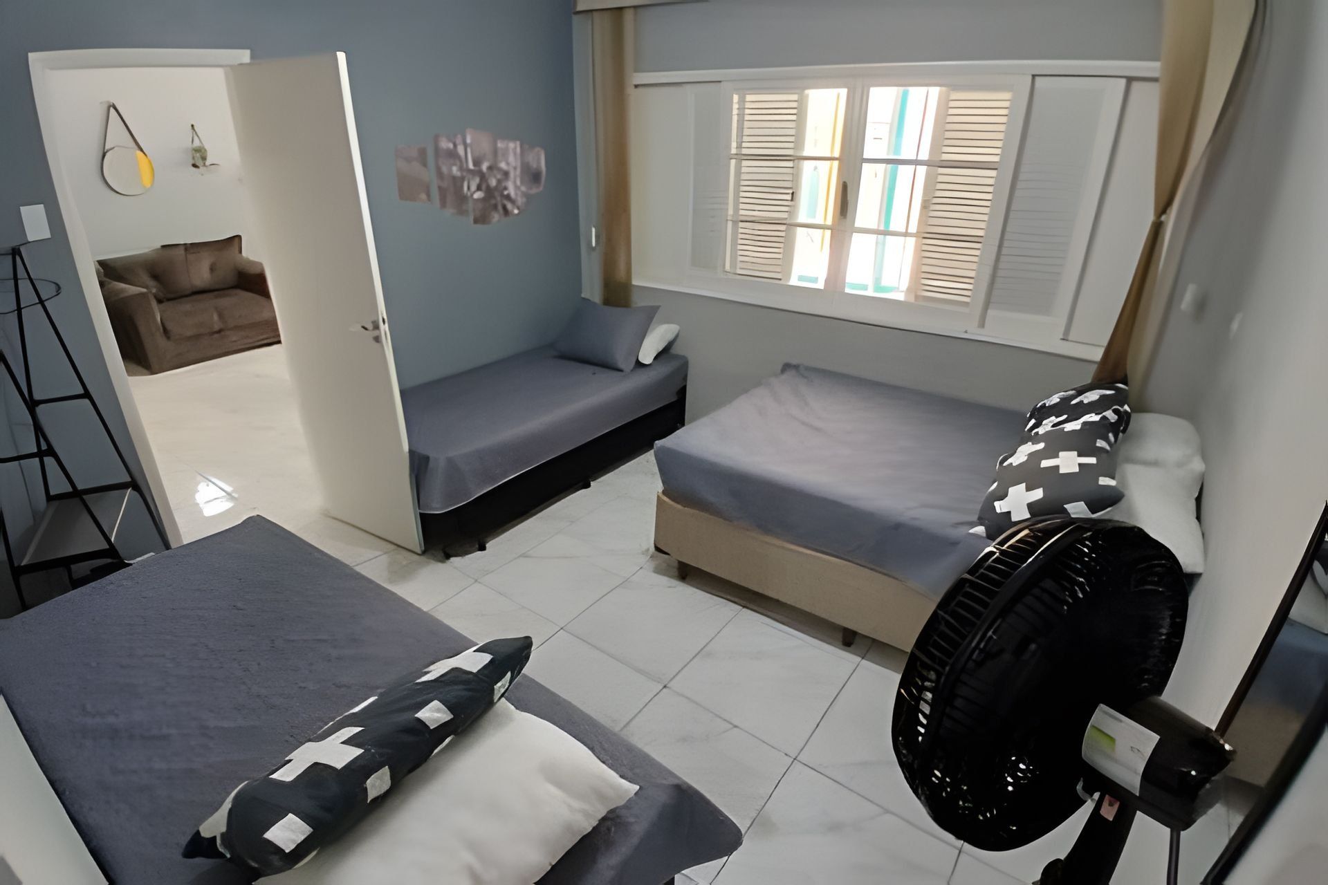 Ein kleines Zimmer mit zwei Einzelbetten, einem kleinen Schlafsofa, einem Standventilator und einem Fenster mit Rollläden.