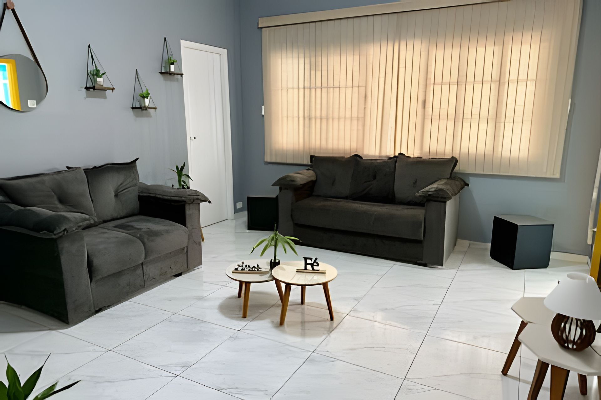 A modern living room with a grey sofa, two small tables, a potted plant, and a large window with blinds.
