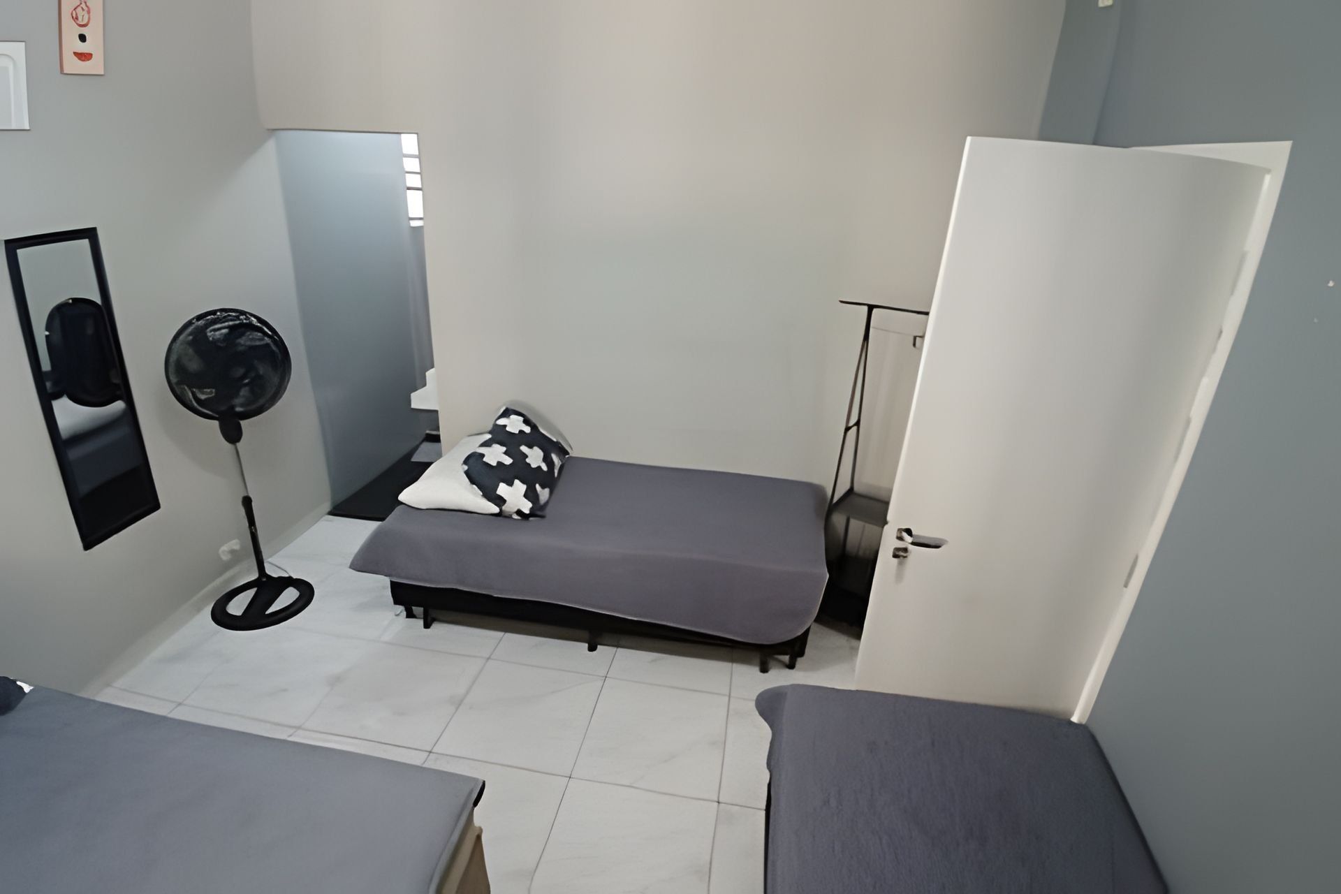 A small, compact room with a single bed, a bedside table, a standing fan, and a door leading to another room.