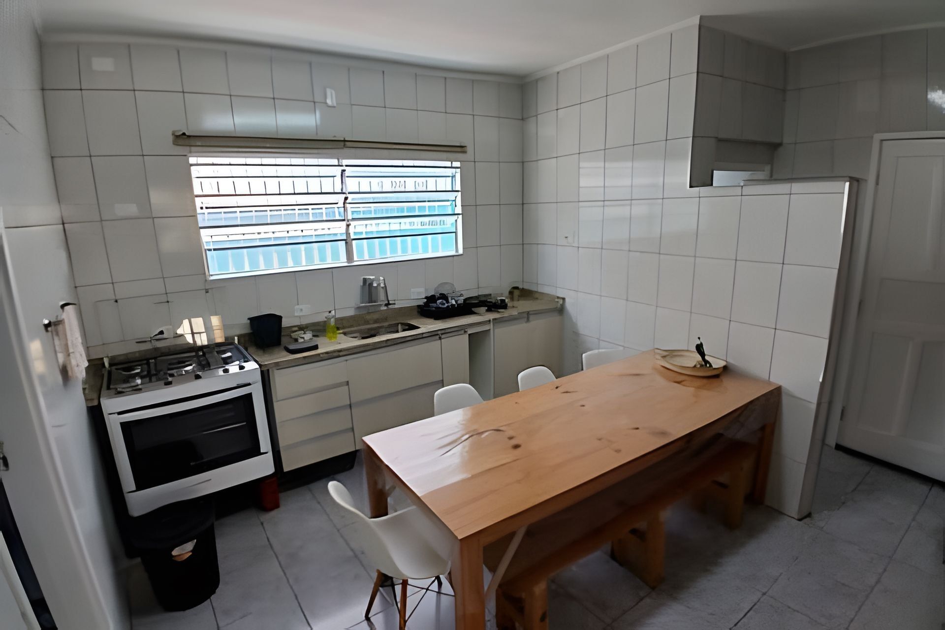 A compact kitchen with white tiles, a window, a stove, a sink, and a wooden dining table with chairs.