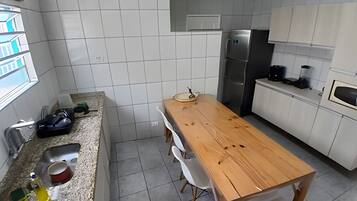 Una cocina con paredes de azulejos blancos, una mesa de madera, sillas blancas, un refrigerador de acero inoxidable y un microondas.