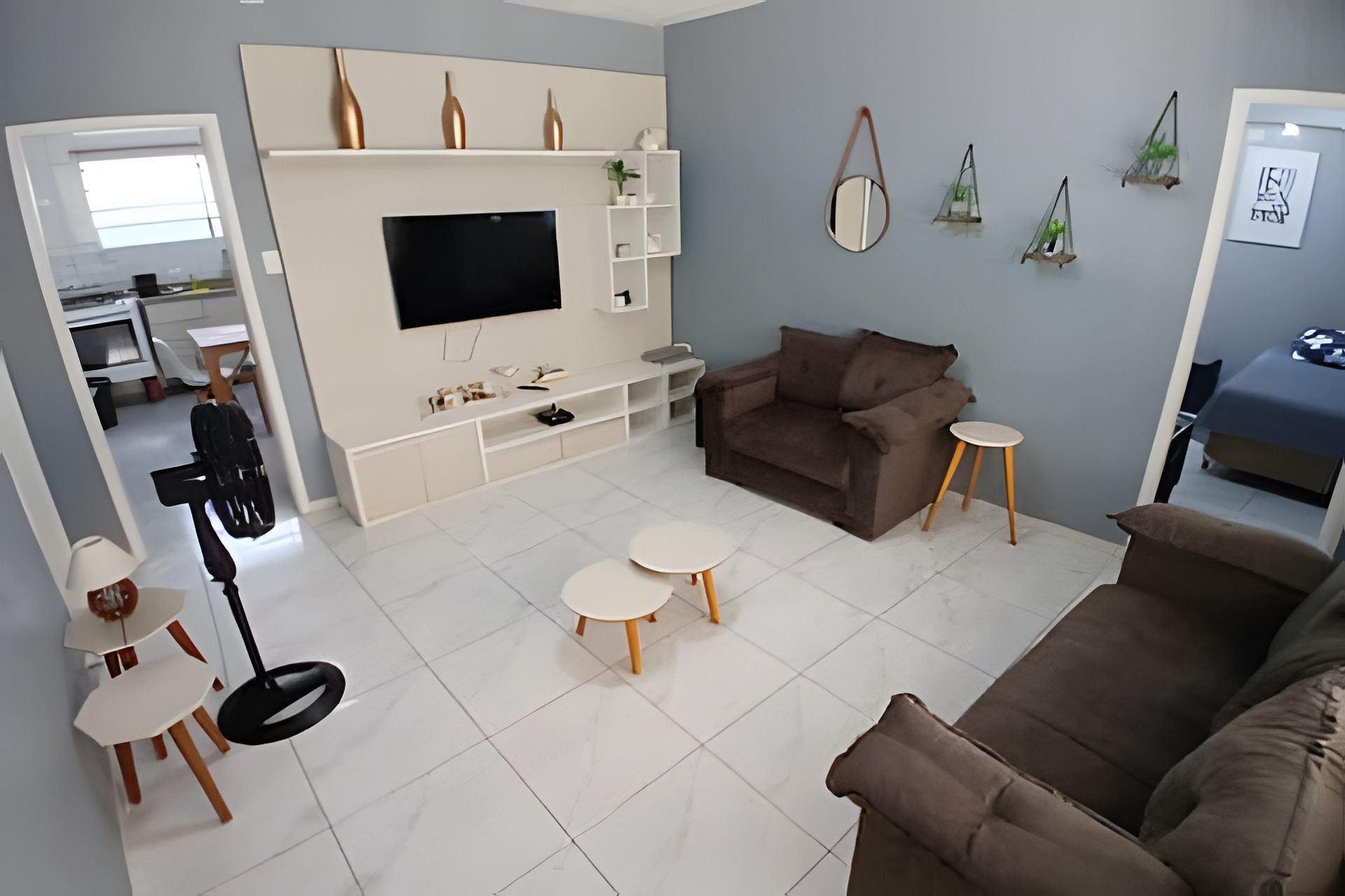 Uma sala de estar moderna com uma televisão de tela grande, um sofá marrom, duas mesinhas redondas e um móvel de entretenimento branco com objetos decorativos.