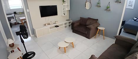 A modern living room with a large flat-screen TV, a brown sofa, two small round tables, and a white entertainment unit with decorative items.
