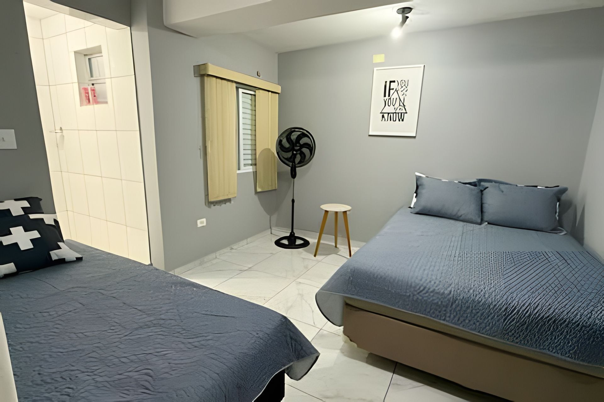Un dormitorio con dos camas, un ventilador de pie, un taburete pequeño y una cita motivacional enmarcada en la pared.