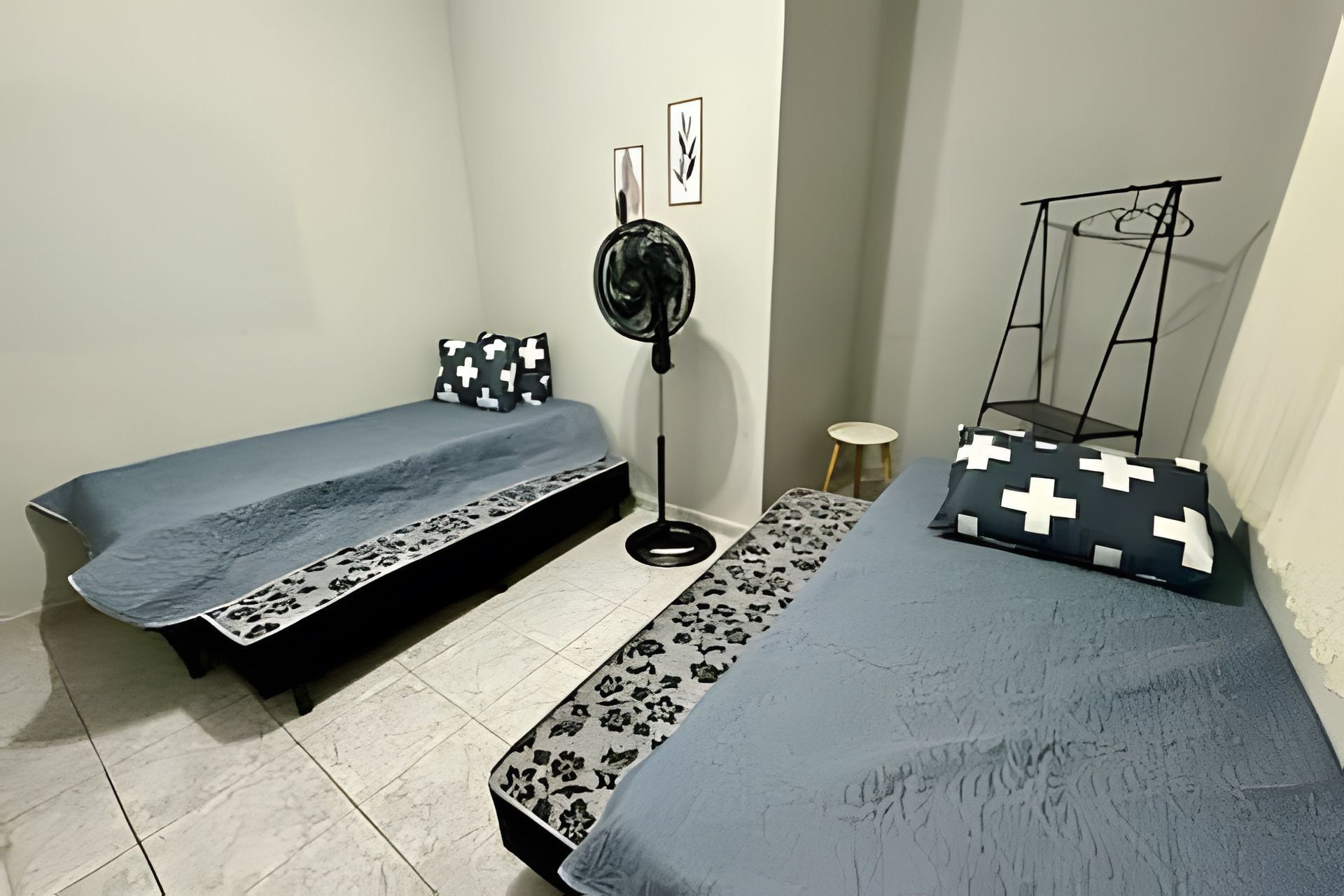 Una habitación pequeña y funcional con dos camas individuales, un ventilador de pie, una mesita y una silla.