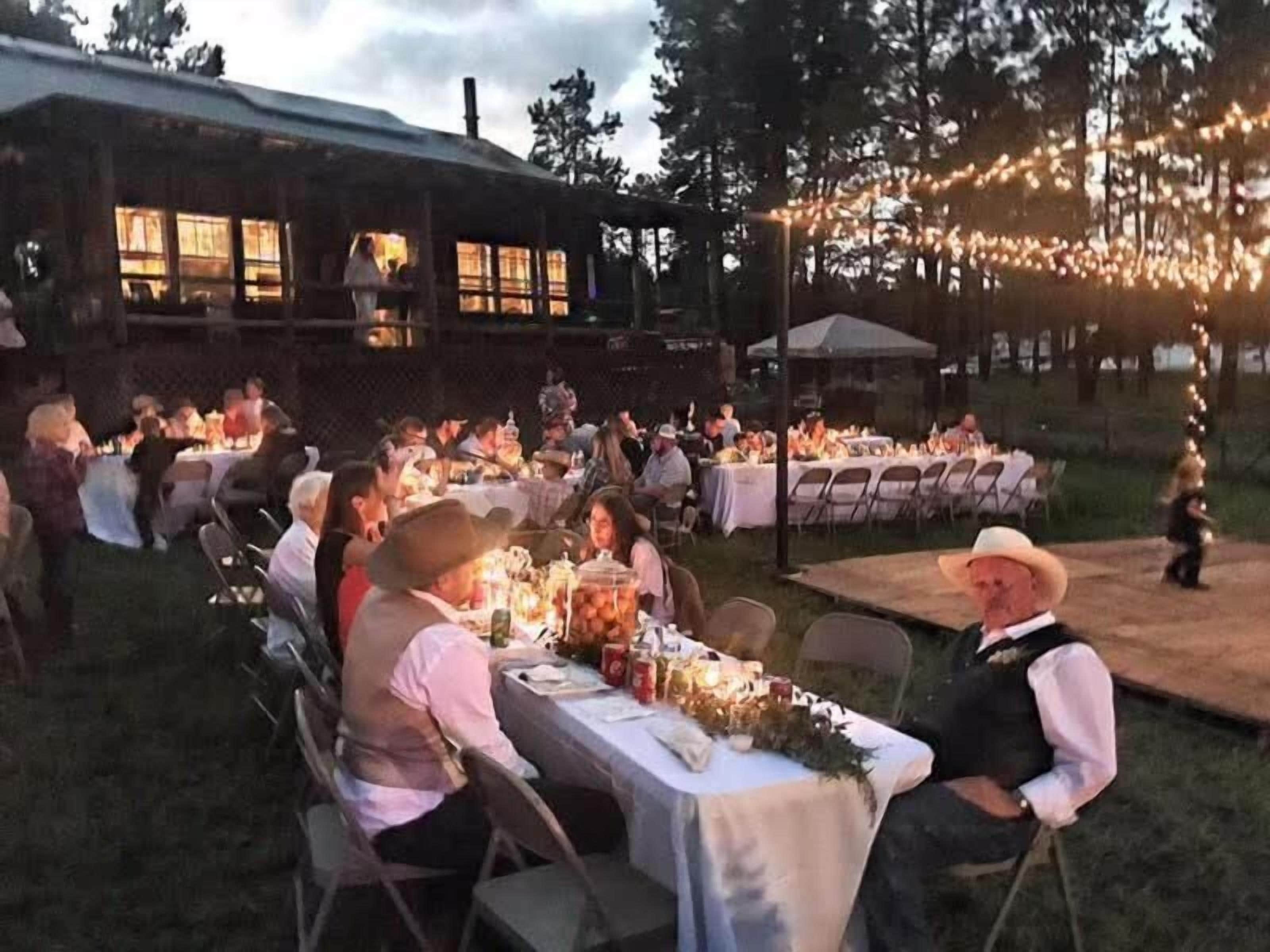 Un espacio exterior para comer con mesas dispuestas para una comida, rodeado por un edificio rústico y luces colgantes.