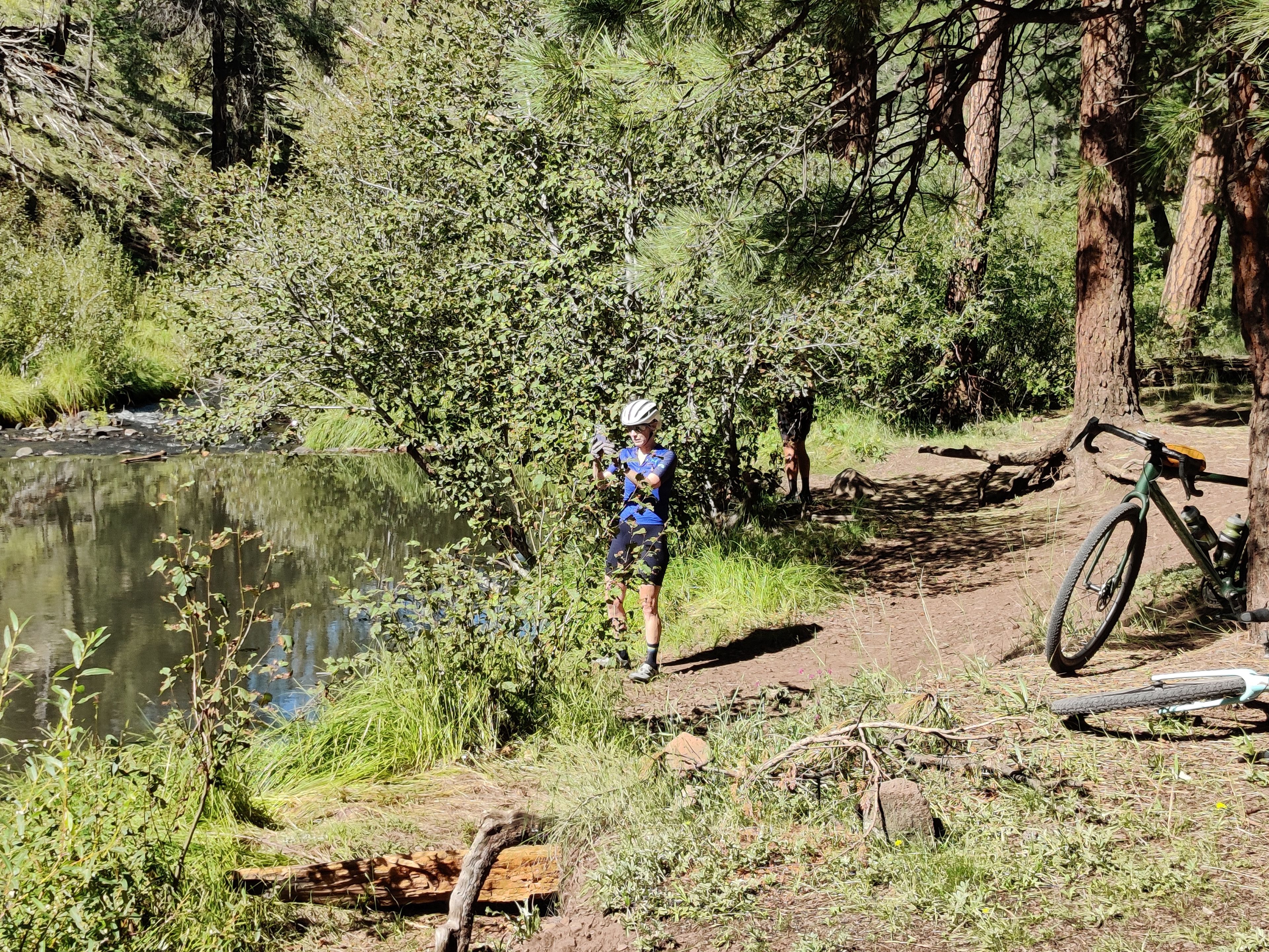 A person standing by a river with a bicycle leaning against a tree.