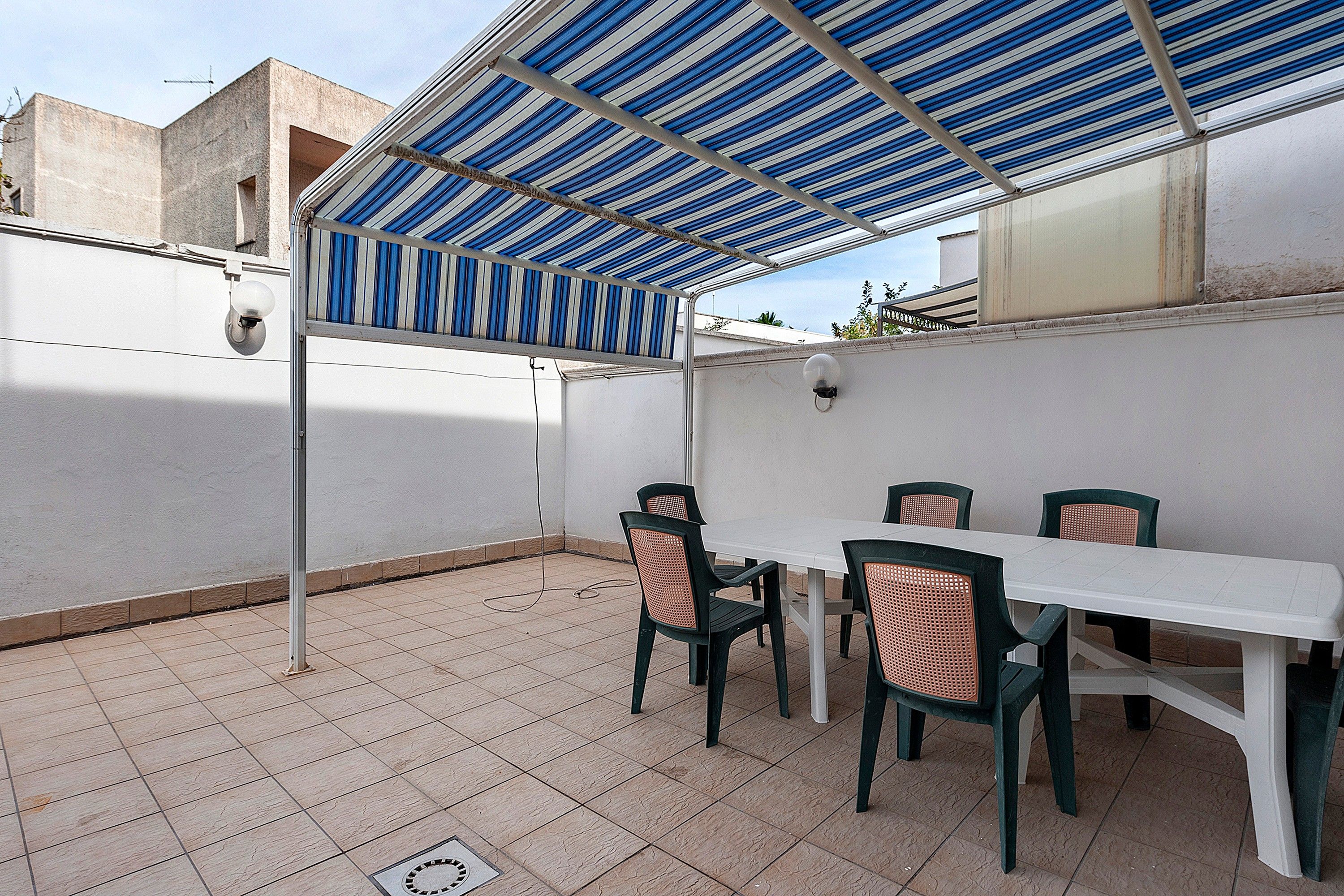Een overdekt terras met een witte tafel en stoelen, een metalen hekwerk en een betegelde vloer.