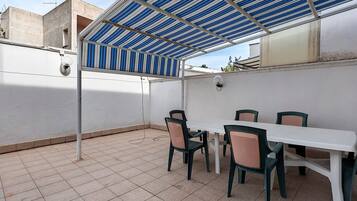 Een overdekt terras met een witte tafel en stoelen, een metalen hekwerk en een betegelde vloer.