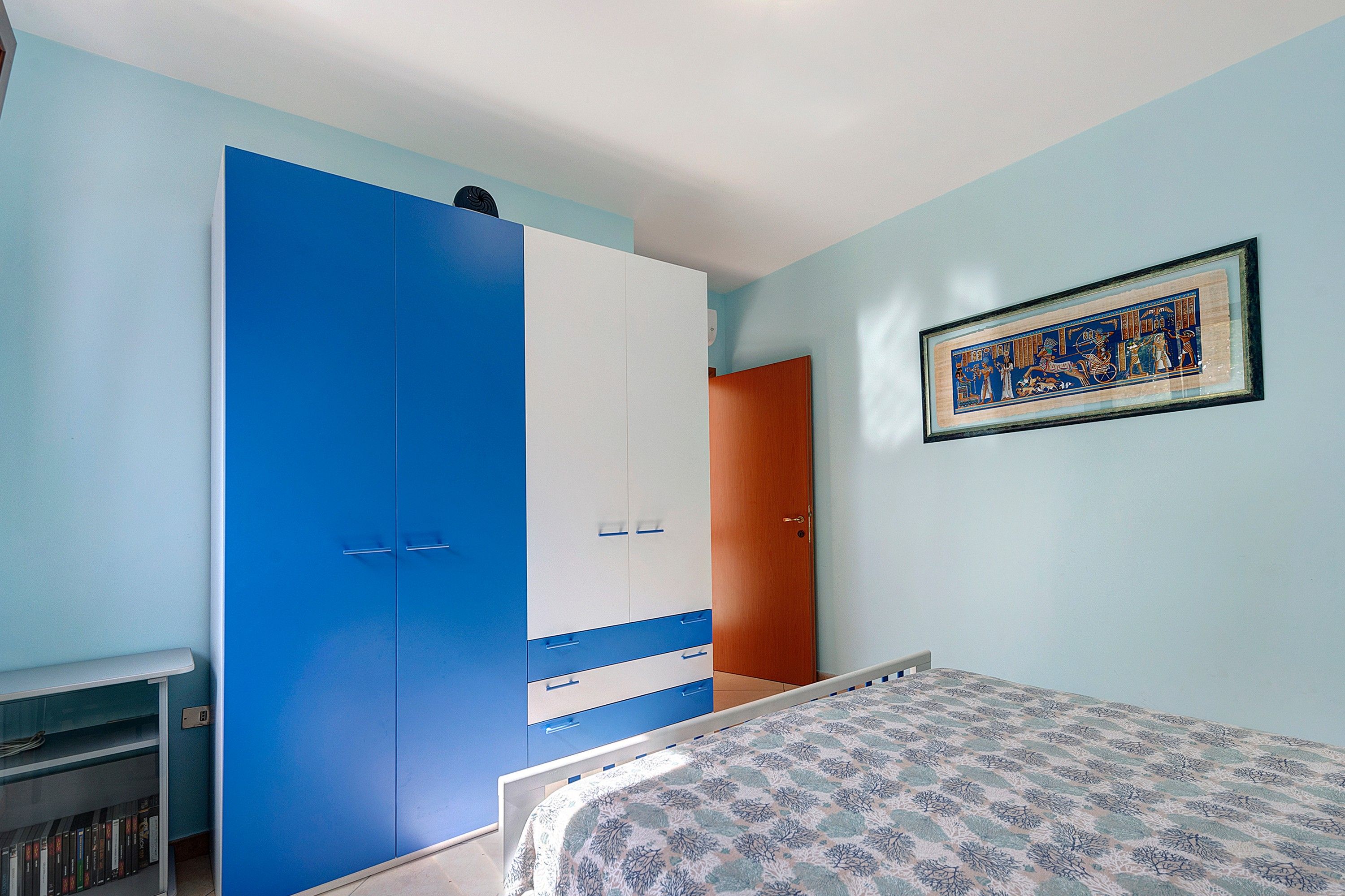Een slaapkamer met een bed, een kledingkast, een deur en een ingelijste foto aan de muur.