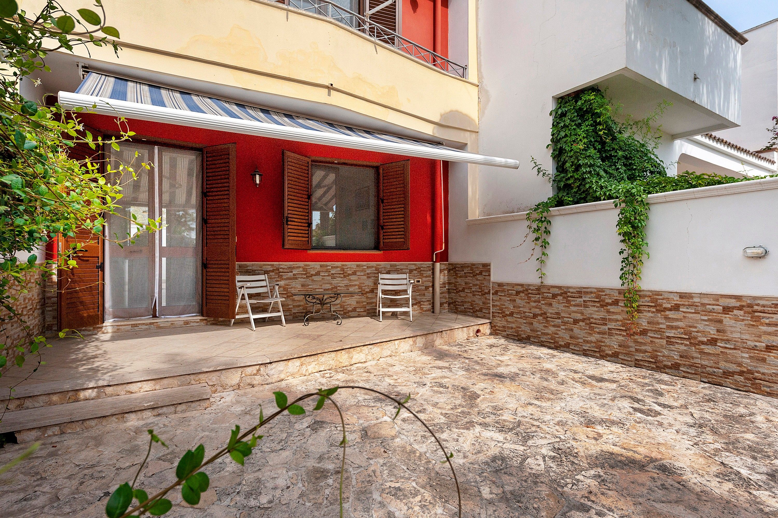 Un edifici residencial amb façana vermella, un balcó i una petita zona de pati amb dues cadires i una taula.