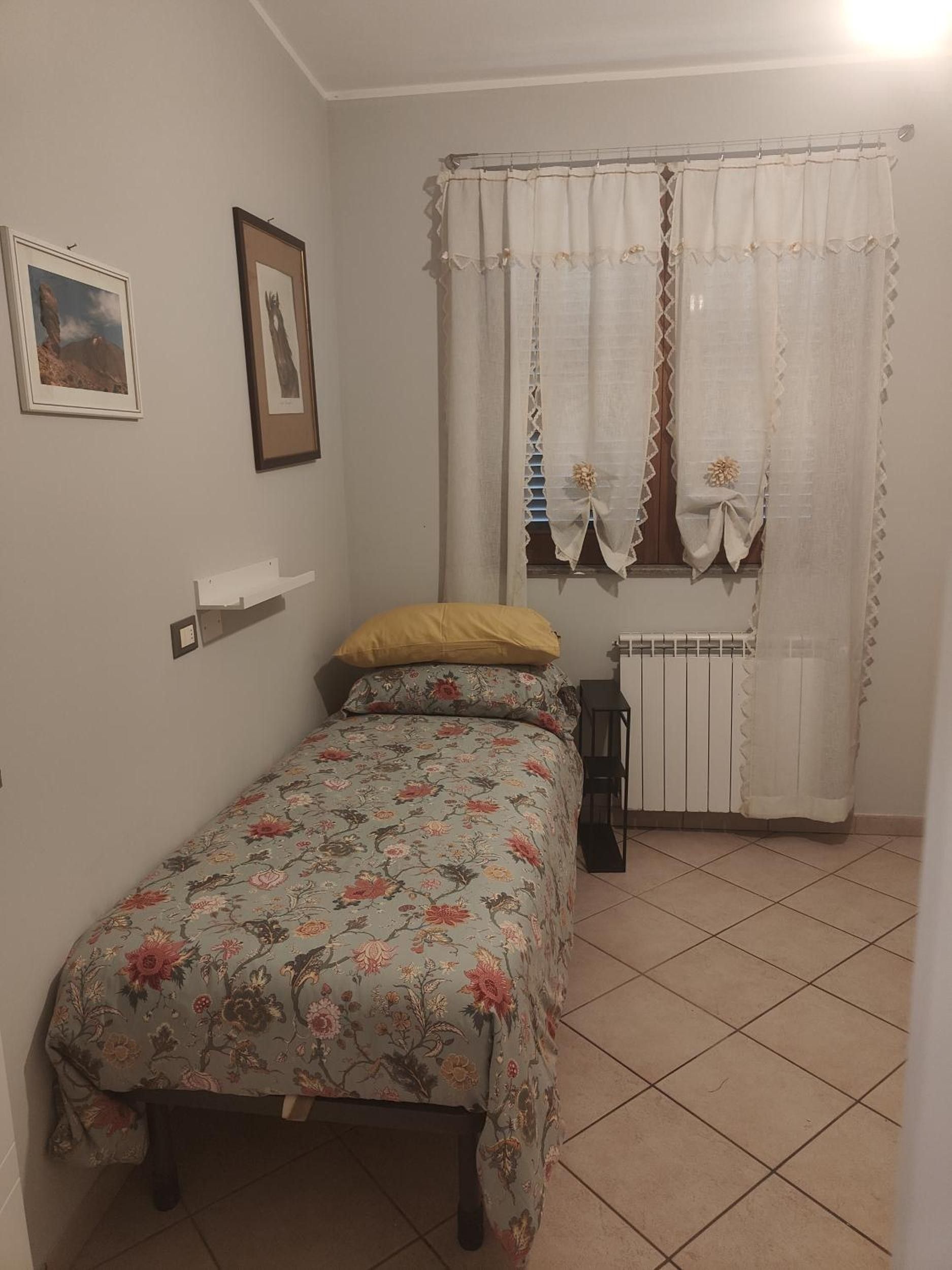Une chambre à coucher avec un lit, un radiateur, une fenêtre avec des rideaux et des tableaux accrochés au mur.
