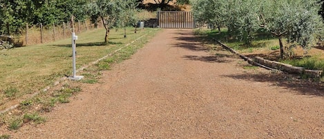 Uma trilha de cascalho leva a um edifício com portão de metal, cercado por árvores e arbustos.