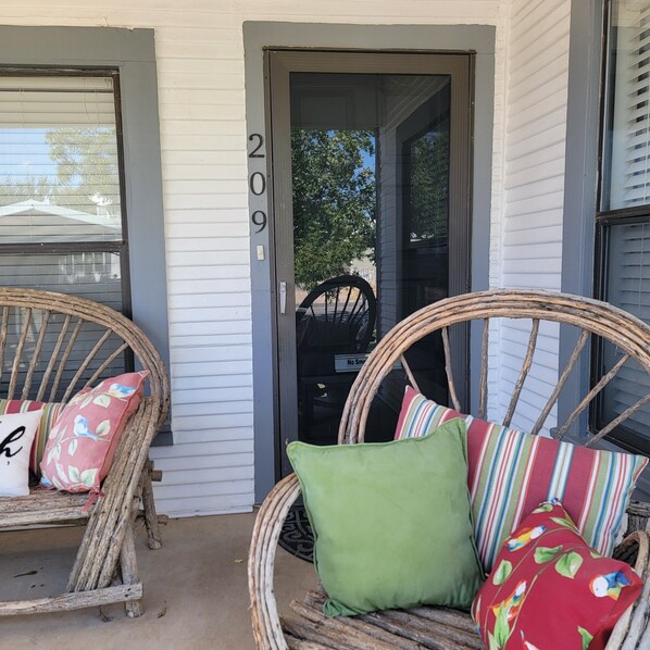 A porch with wicker chairs and cushions, a door with the number 209, and a view of the street and trees.