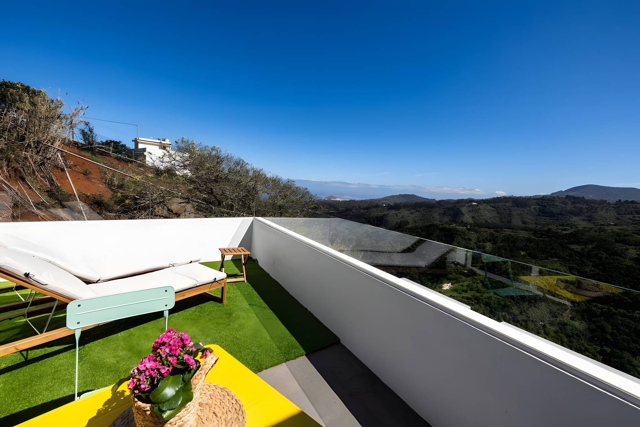 Un balcón con vistas a un paisaje montañoso, una banca de madera y una pequeña mesa con un jarrón de flores.