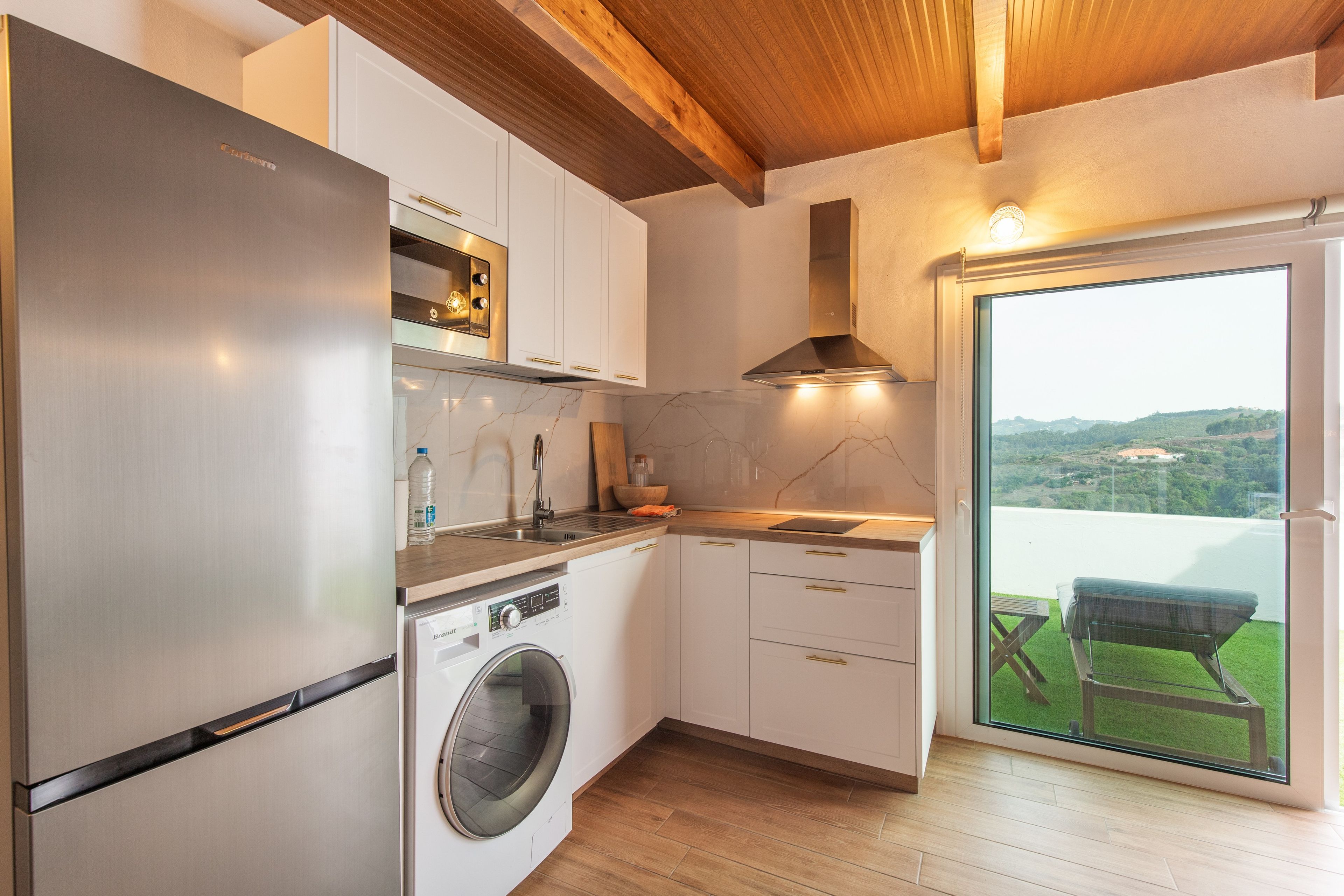 A modern kitchen with a built-in washing machine, a large window, and a view of a green landscape.