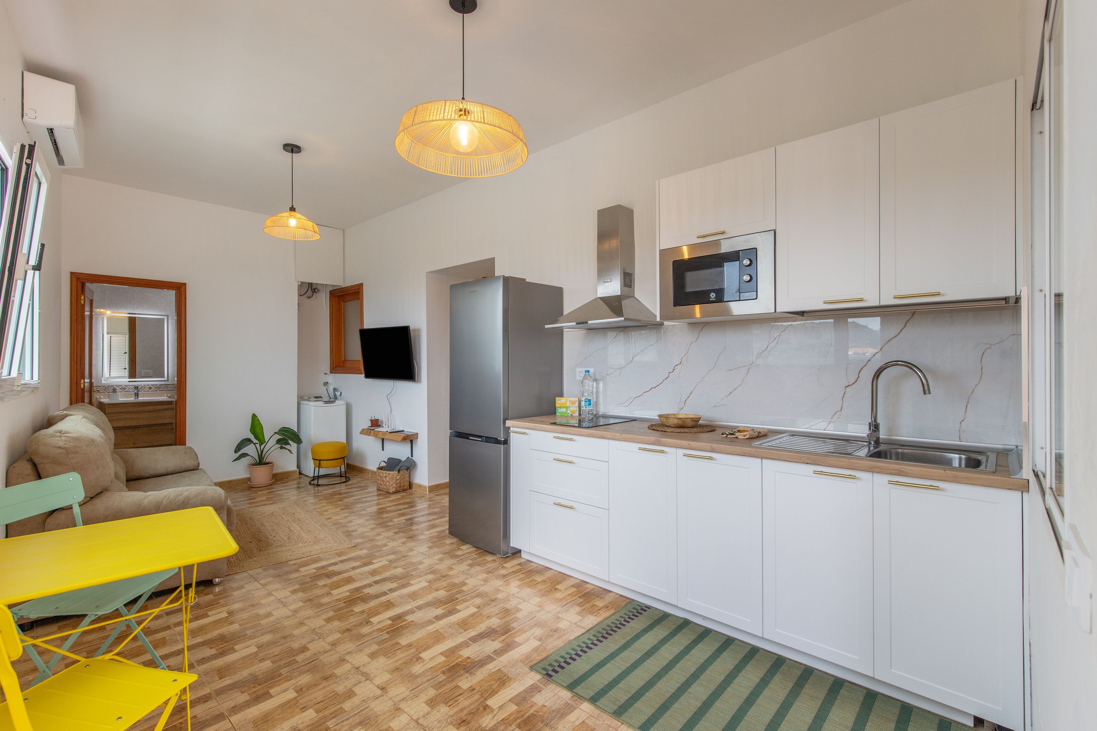 Una cocina moderna con armarios blancos, un refrigerador de acero inoxidable y un microondas. Hay una mesa de comedor de madera y una silla amarilla en la zona de estar.