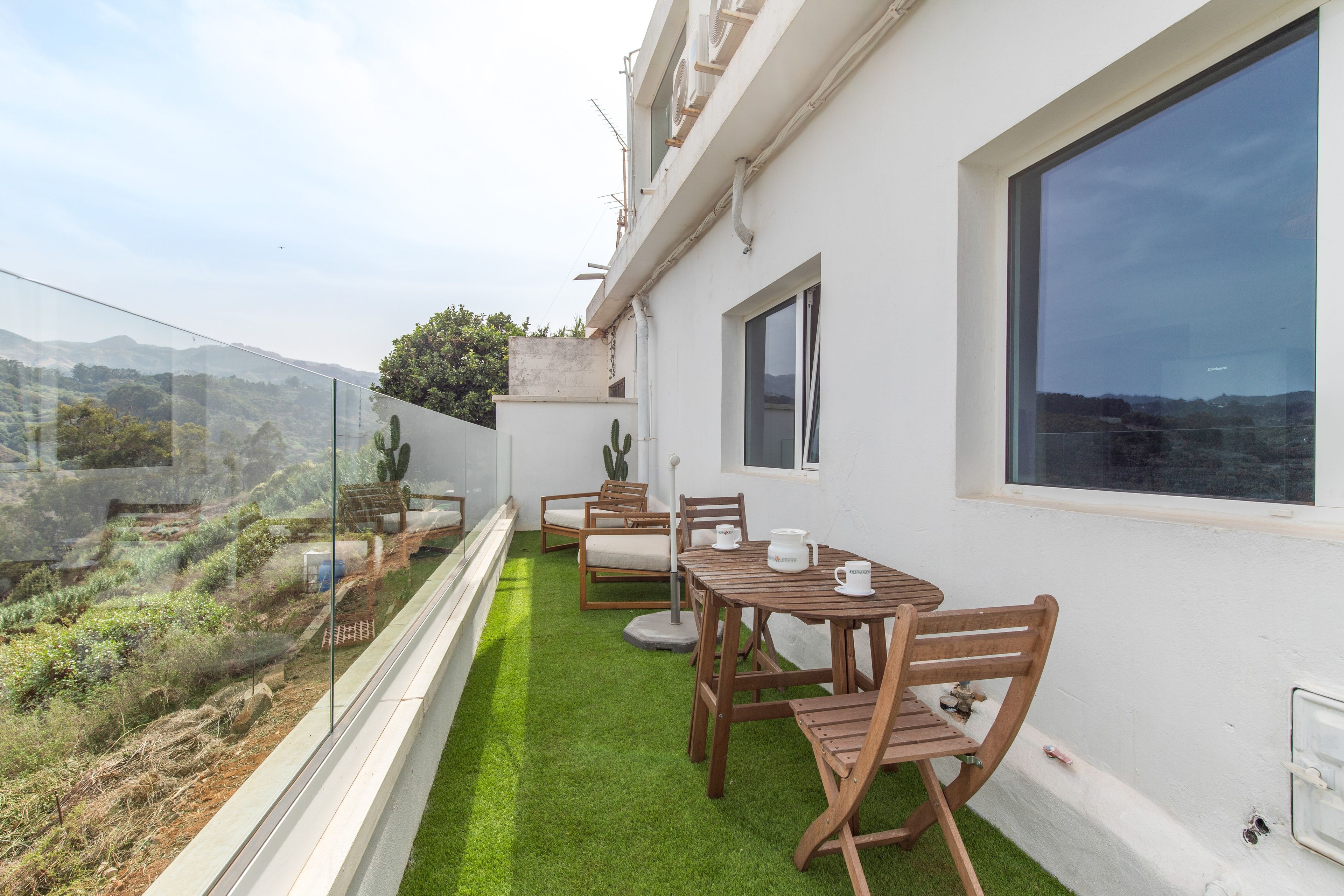 A balcony with a glass railing, wooden furniture, and a view of the landscape.