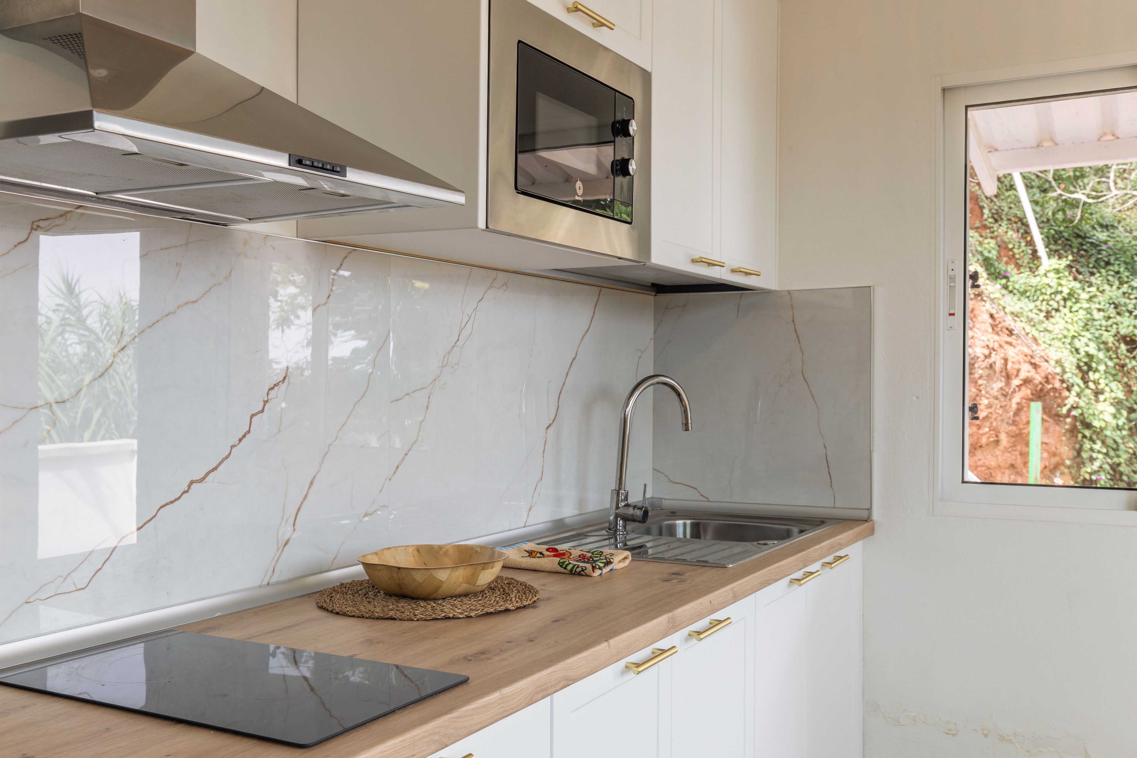 Une cuisine avec des meubles blancs, un évier en acier inoxydable, un plan de travail en bois et une fenêtre donnant sur un paysage verdoyant.