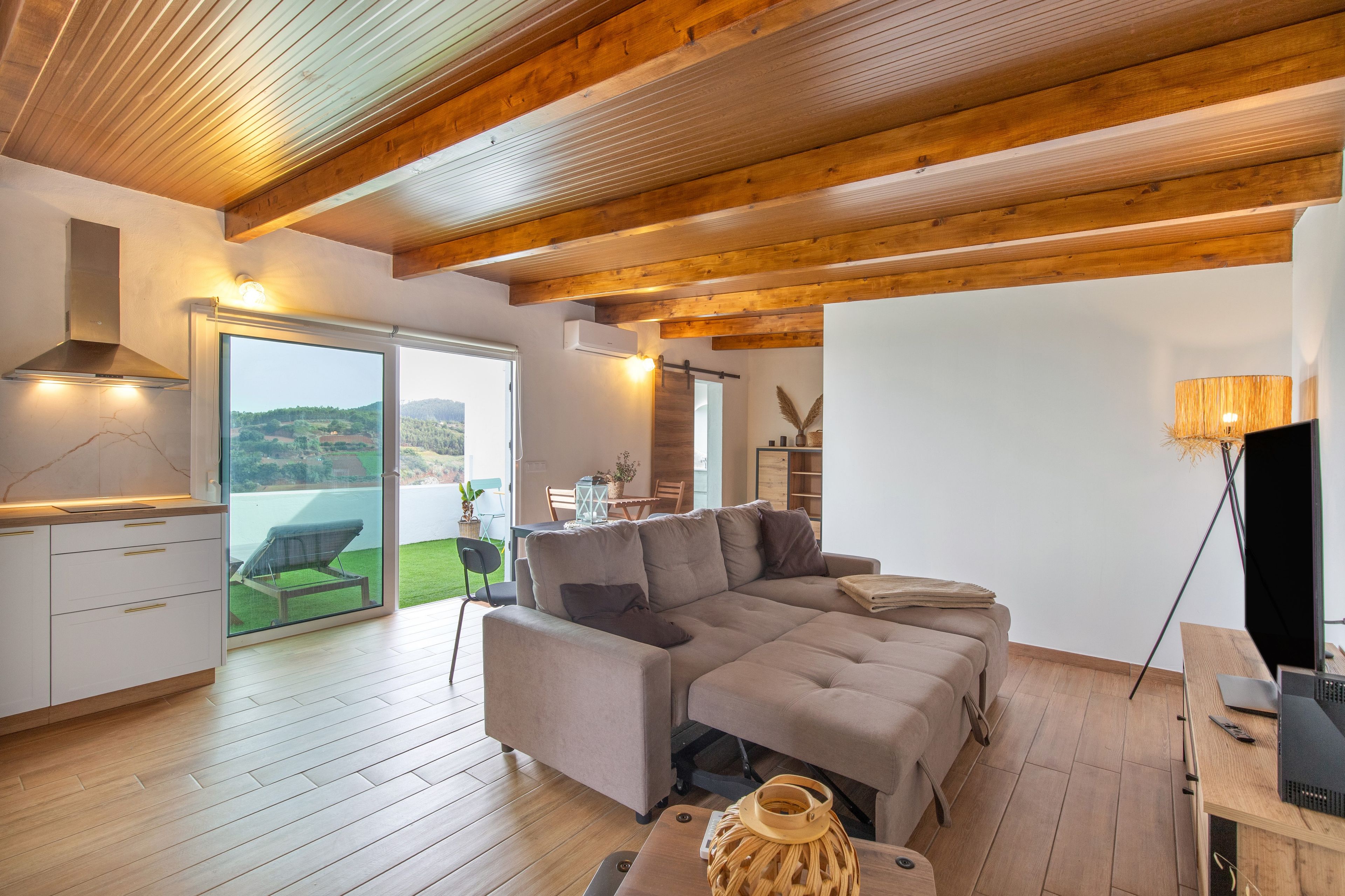 Ein modernes Wohnzimmer mit Holzboden, einem grauen Sofa, einem Essbereich und Blick auf eine grüne Landschaft durch große Fenster.