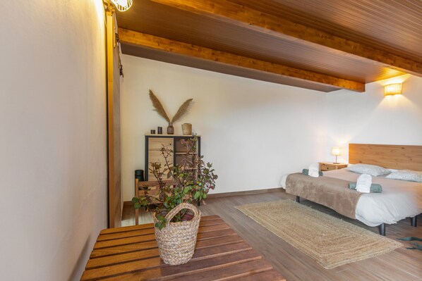 A bedroom with a wooden ceiling, a bed with white bedding, a bedside table with a lamp, a woven basket with plants, and a wall-mounted shelf with decorative items.