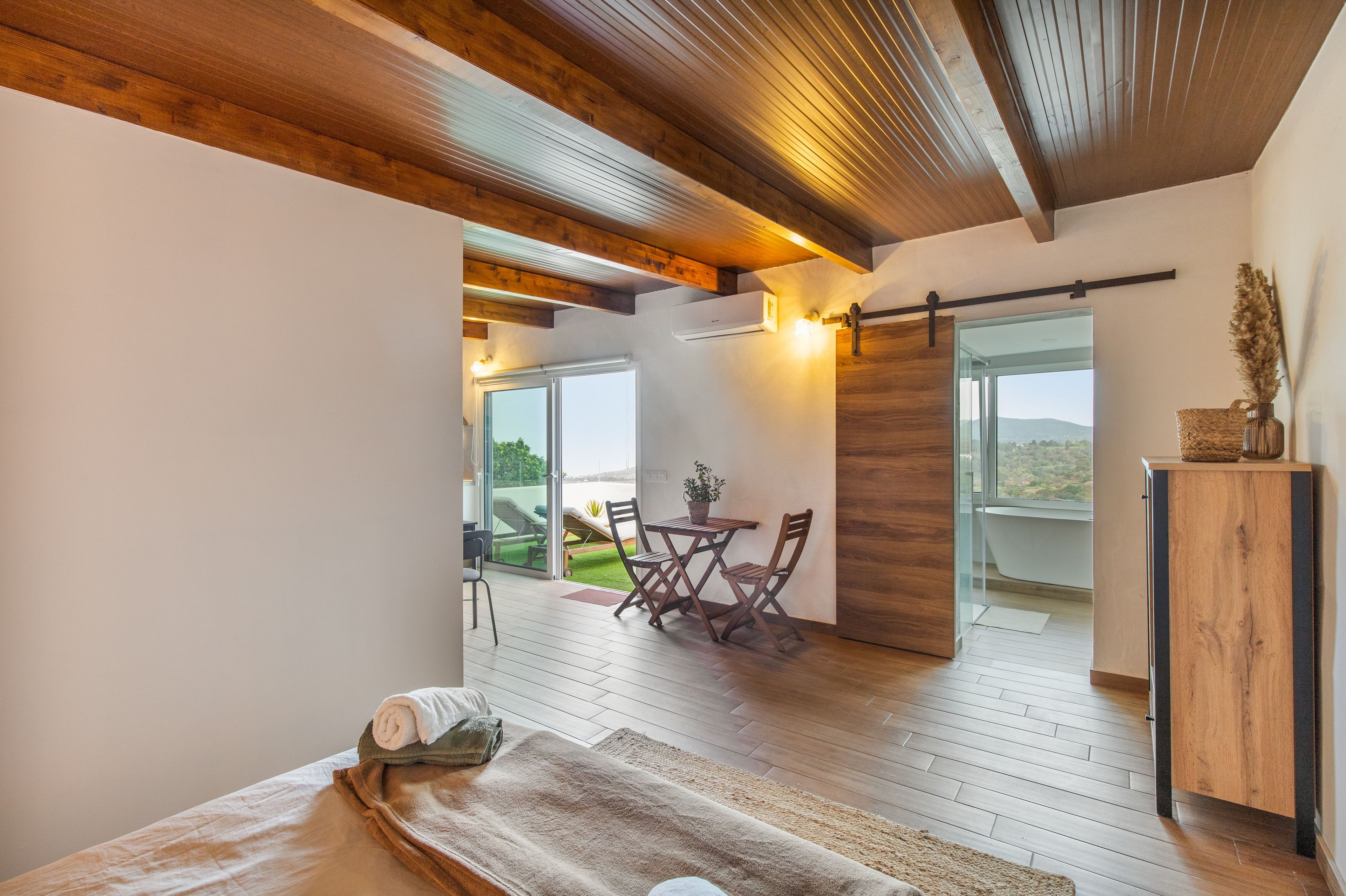 Une chambre moderne avec un grand lit, un plafond en bois, une vue sur l’extérieur et une petite table avec des chaises.