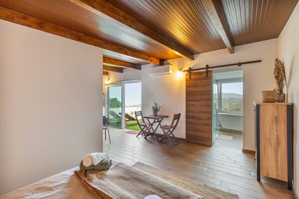 A modern bedroom with a large bed, a wooden ceiling, a view of the outdoors, and a small table with chairs.